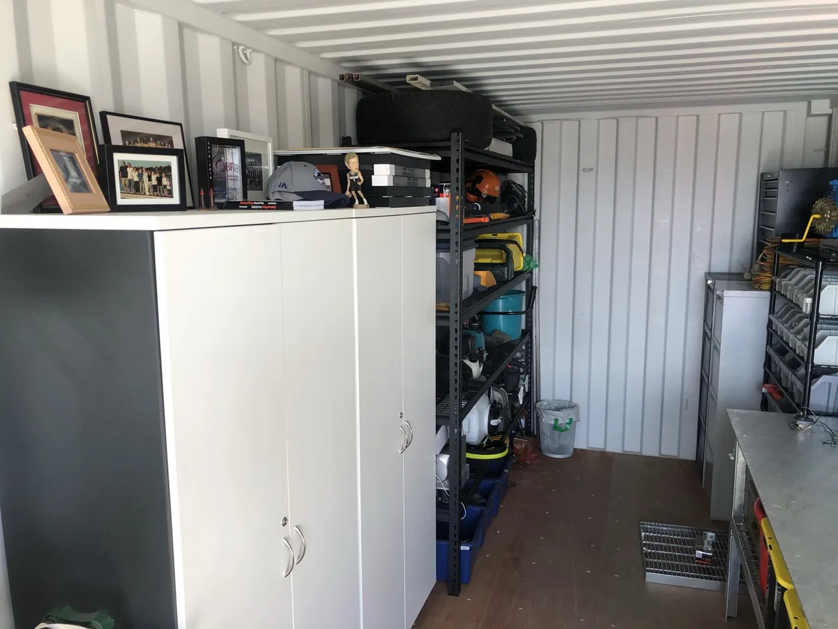 The other side of Ned's container: tall white cupboards with framed team photos on top, and black shelves of garden and power tools