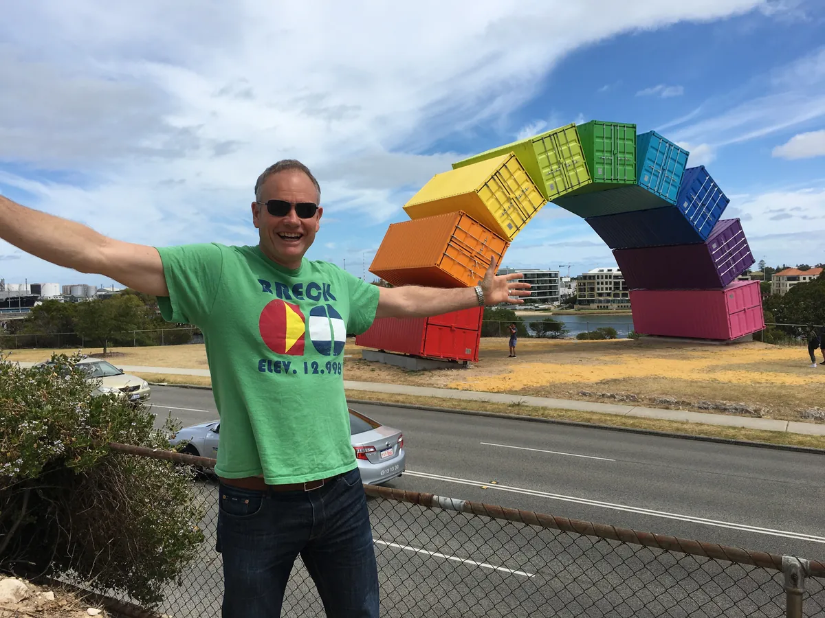 Ned Coten with his arms out in front of a rainbow arch made of brightly painted shipping containers